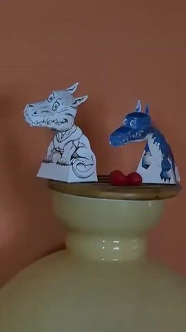 The video displays two paper dragon cutouts placed on top of a yellowish ceramic object, likely a vase or a lamp base. The dragon on the left is monochrome with intricate drawings of scales and clothing. The dragon on the right is colored blue and white, with visible wings. Two small red objects, possibly berries or candy, are situated between the two dragons. The background is an orange wall with a white electrical outlet visible on the left side of the frame. The lighting is warm and casts soft shadows. The camera is positioned at eye level with the dragons, and the shot remains static throughout the brief clip.