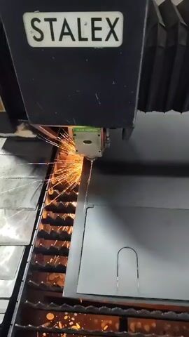 A close-up shot shows a STALEX brand laser cutting machine in operation. The machine's head is positioned over a large, flat sheet of gray metal. A bright, focused beam of light emanates from the cutting head and is actively cutting through the metal. Sparks and molten material are seen flying downwards as the laser severs the material. The metal sheet has pre-cut lines visible, indicating a pattern is being followed. The cutting head moves along these lines, creating precise cuts. The background shows a dark industrial setting with the machine's internal structure visible, including a gridded support surface below the metal sheet where the sparks are falling. The overall impression is one of precise, industrial manufacturing.