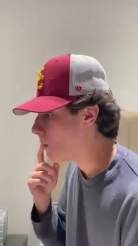 A young man wearing a USC baseball cap and a gray long-sleeved shirt is seen in profile, exhaling a puff of vapor, possibly from a vape or e-cigarette. He is holding a finger to his chin in a pensive manner. In the background, a partially visible beer bottle stands on a surface near electrical outlets and a beige towel. The setting appears to be indoors, against a light-colored wall.