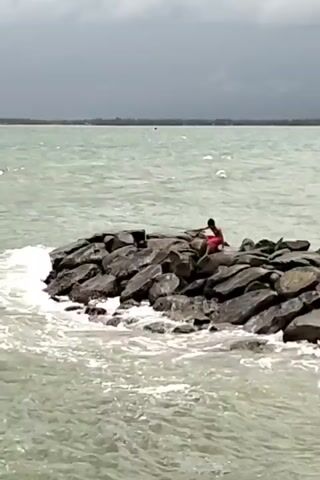 The video shows a person sitting on a rock formation in the ocean. The person is wearing a red shirt and appears to be fishing. The waves are crashing against the rocks, and the sky is overcast. The water is choppy and green. In the background, there is a strip of land.