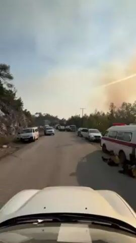 Here is a detailed description of the video:

The video shows a white car driving down a road lined with parked vehicles, with smoke visible in the distance, likely from a fire. The road is surrounded by trees and a rocky hillside. Several types of vehicles can be seen parked along the road, including cars, vans, an ambulance, and a small tanker truck. People can be seen standing next to some of the vehicles. The sky is partially cloudy, with a haze from the smoke. The video is shot from the perspective of the driver of the white car.