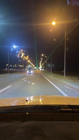 The video shows a view from inside a vehicle at night, looking out through the windshield onto a highway. Streetlights illuminate the road, and there are several vehicles visible, including a large truck in the distance. Green and purple laser beams are visible crossing the road in front of the vehicle. The windshield has some water droplets on it, causing some glare and distortion of the lights.