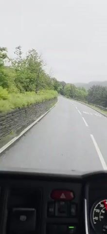 The video is a first-person perspective shot from the dashboard of a vehicle, likely a bus or truck, driving on a paved road on a cloudy day. The road curves gently to the left, with a stone wall and lush green foliage lining the left side. Trees and hills are visible in the distance on both sides of the road. White dashed lines mark the lanes of the road. The dashboard of the vehicle is visible at the bottom of the frame, with a red hazard light button and parts of gauges. The audio features the sound of a vehicle engine and tires on a wet road, and a faint snippet of what sounds like music.