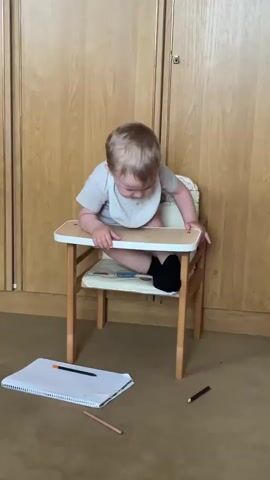 A toddler sits in a small, wooden high chair. The toddler wears a light-colored t-shirt and a white bib. The high chair has a white tray with a light-colored cushion on the seat. The toddler is looking down at the tray, with their right leg bent and tucked under them, and their left leg extended out. On the floor in front of the high chair is a spiral-bound notebook, open to a blank page. Two pencils lie next to the notebook, one orange and one light brown. A dark brown pencil lies on the floor to the right of the high chair. The background is dominated by a wooden wardrobe with two doors. The floor is carpeted in a neutral color. The scene appears to be indoors, likely in a home, given the presence of a wardrobe and a child's high chair. The lighting suggests it could be daytime. The child appears focused on something on the tray or the notebook.