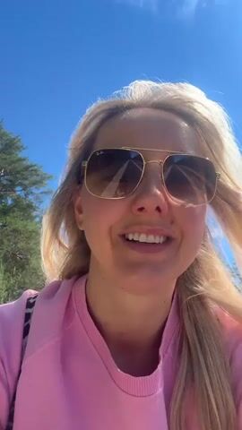 This media file captures a moment of outdoor activity, likely a bike ride, in Carnikava, Latvia. The primary subject is a woman with blonde hair, wearing large aviator sunglasses, a bright pink sweatshirt, and red shorts. She is smiling broadly at the camera, suggesting a cheerful and engaged mood. She is on a bicycle, with her right hand on the handlebars.

Behind her, a young boy is also riding a bicycle. He is wearing a helmet, a blue tie-dye t-shirt, and grey shorts, and has a backpack on. He appears to be looking ahead as he cycles. Both subjects are on a paved path, with a road visible in the background. The surrounding environment includes trees, suggesting a natural or park-like setting, possibly near a forest given the presence of tall pines. The sky is a clear, vibrant blue, indicating fair weather and a sunny day. The lighting suggests it is daytime, likely mid-morning or afternoon due to the bright sunlight and the distinct shadows cast by the bicycles.

The overall scene is one of leisure and recreation, with the woman and child enjoying an outing together. The bright colors of their clothing and the sunny weather contribute to a positive and energetic atmosphere.