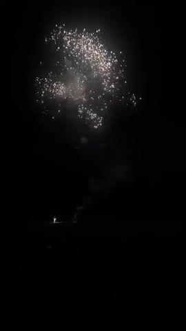 A fireworks display is occurring against a black night sky. Sparks and bursts of light are visible, with smoke trailing from some of the explosions. In the lower portion of the frame, a dark, flat landscape is visible with a few faint lights, possibly indicating people or small structures. One bright light, possibly a person holding a light source or a small firework, is visible in the lower left. Another fainter light is visible to the right of that, and a small reddish-orange light appears to be a firework being ignited or burning on the ground. The fireworks are ascending and exploding in the sky.
