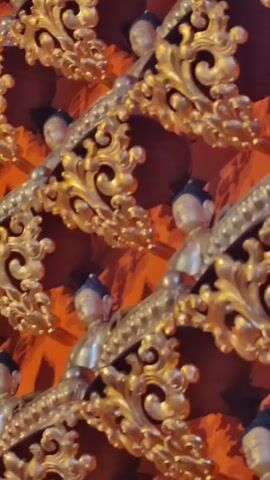 The video pans across a wall decorated with a large number of small, golden Buddha statues. The statues are arranged in rows, each placed on a decorative golden shelf. The wall behind the statues is a vibrant orange color. The overall impression is one of religious art and devotion.