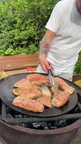 A person wearing a white t-shirt and white shorts is grilling salmon on a large, round, flat griddle over an open fire pit in an outdoor setting. The griddle is placed on a metal ring filled with glowing charcoal. Several pieces of seasoned salmon are arranged on the griddle, some of which are being flipped by the person using metal tongs. The person has tattoos on their left arm.

The scene is set outdoors with lush green foliage in the background, suggesting a sunny day, possibly late afternoon or early evening, given the natural lighting. A wooden bench is visible behind the person. The environment appears to be a backyard or garden area. There is no visible text in the media. The overall mood is one of casual outdoor cooking and enjoyment. The location is Rezekne, Latvia.