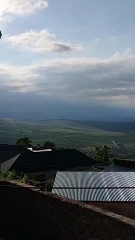 The video shows a landscape view from a high vantage point, looking down on a valley with rolling green hills and a distant town or village. The sky is partly cloudy with some sun breaking through. In the foreground, several rooftops are visible, made of corrugated metal and darker colored materials, suggesting a rural or semi-rural setting. A stone wall runs along the bottom edge of the frame. There are no people or obvious actions taking place in the video.