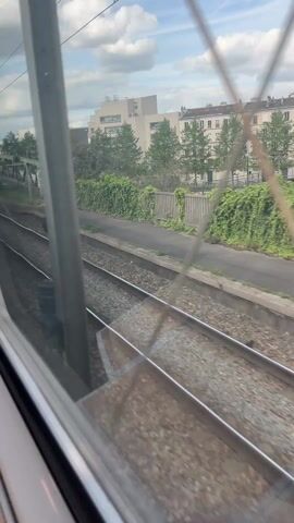The video shows a view from inside a moving train, looking out the window at the passing scenery. The main focus is on the railway tracks running parallel to the train. Beyond the tracks, there's a green metal bridge spanning over a road. Buildings and greenery are visible in the background under a cloudy sky. Graffiti covers a wall alongside the tracks. The camera seems to be moving at a moderate speed, judging by the pace of the passing objects. The scene captures a typical urban or suburban landscape viewed from a moving train.