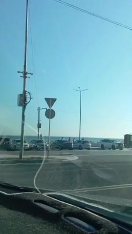 The media file shows a daytime view from a vehicle moving along a coastal road. The sky is clear and blue, suggesting good weather. The scene features a beach with people visible in the distance, as well as a road with several cars and a truck. Some of these vehicles appear to be parked, while others are in motion. 

Notable objects include a parking sign with a "P" symbol, a street lamp, and a building with a sign that reads "Golden Beach". The overall atmosphere appears relaxed and leisurely, consistent with a beachside location during the day. The presence of the sea and the beach indicates a coastal setting, and the text on the sign confirms it as "Golden Beach". The time of day is clearly daytime, indicated by the bright sky and clear visibility. The activity taking place is general movement along a road that runs parallel to a beach area, with people enjoying the seaside.