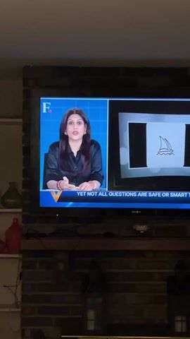 A television displays a news program with a female news anchor with long dark hair. She is wearing a dark blouse and is gesturing with her hands. The screen is divided into two sections, with the anchor on the left and a logo for Microsoft on the right. The caption at the bottom of the screen reads "YET NOT ALL QUESTIONS ARE SAFE OR SMART." The television is mounted on a brick wall, and below the TV is a mantle. The setting appears to be a living room, as indicated by the shelves to the left with various vases and decorative items on them.