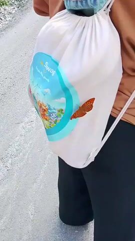 A person wearing a brown shirt and black pants walks on a gravel path. A white drawstring bag with a butterfly image and a blue circular design is slung over their shoulder. A denim piece is tied around the top of the bag. Another person, wearing a red bucket hat, holds the bag with their left hand. The person in the brown shirt looks over their right shoulder.