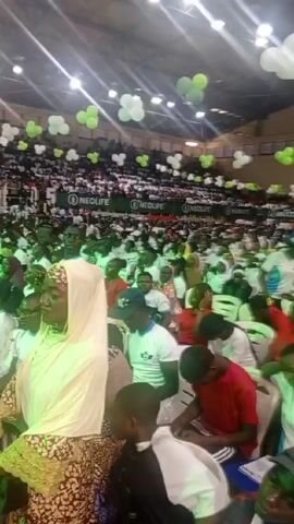 The video shows a crowd of people inside a large hall or auditorium. Many attendees are dressed in white or red shirts, and some are wearing green. They are sitting or standing, with many holding up their phones, seemingly recording or taking pictures. The hall is decorated with green and white balloons in the shape of clovers. In the foreground, people are also holding up phones, capturing the event. The atmosphere appears lively and celebratory.