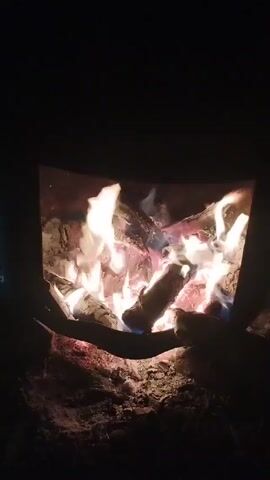 The video shows a close-up of a fire burning inside a metal fire pit or stove. The flames are bright and orange-white, licking around pieces of wood that are partially burnt. Embers glow red beneath the flames. The surrounding area is dark. The camera zooms in slightly, focusing on the center of the fire.