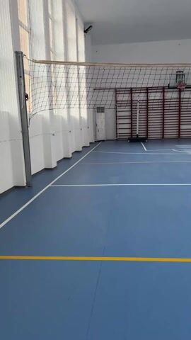 The video shows an indoor volleyball court with a blue floor and yellow lines. Two people are present: one in a black shirt and another in a green shirt. They are on one side of the net. The person in black is bouncing a volleyball. The environment includes wall bars, a basketball hoop, windows, and a volleyball net. Sunlight streams through the windows. The camera slowly pans to the right.
