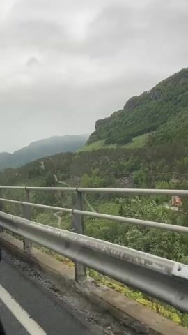 This video captures a drive through a scenic mountain landscape in Skare, Norway. The camera is positioned inside a vehicle, looking out over the side of a bridge or overpass. The foreground features a metal guardrail and a portion of the road, with white lane markings visible. Beyond the railing, a lush green valley unfolds, densely populated with trees. The slopes of a large, verdant mountain dominate the background, its peak partially obscured by a cloudy, overcast sky. The overall atmosphere suggests a cool, perhaps damp, day, typical of a northern climate. The thick tree cover and imposing mountain suggest a remote, natural setting. There are no people or animals visible, and the primary focus is on the natural scenery and the passage through it by vehicle. The time of day is not clearly discernible due to the overcast conditions, but the light appears to be diffused, consistent with daytime. The emotional tone is one of quiet observation of nature's grandeur. No text is visible in the media.