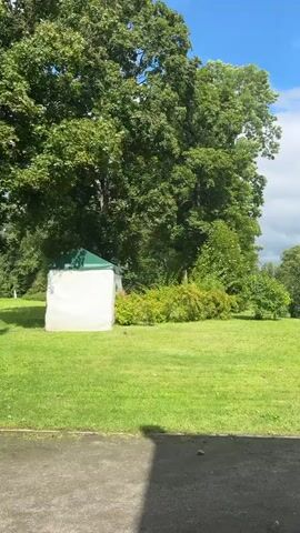 A video shows a park on a sunny day with a few clouds. The camera pans from left to right, revealing a white tent with a green roof on the left side of the frame, surrounded by lush green grass and trees. The camera then moves across a grassy field, showcasing various trees and bushes. The sky is blue with white, puffy clouds. In the foreground, a dark asphalt path with a puddle of water is visible.