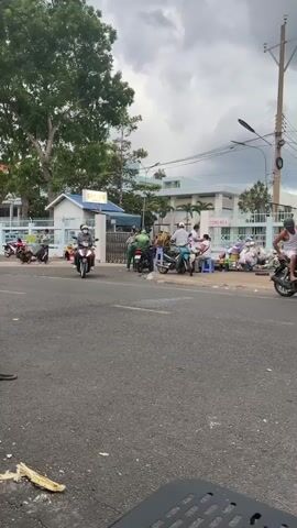The video shows a street scene in Vietnam. The road is asphalt and appears to be in good condition. There are several motorcycles driving on the road, some with multiple passengers. On the side of the road, there is a group of people sitting and standing around a stall selling what appear to be bags of produce. There are also several motorcycles parked near the stall. Behind the stall, there is a building with a sign that says "CONG SO 2". The sky is cloudy and overcast. There is a large tree on the left side of the video. The video appears to have been taken from a stationary position, possibly from a table or chair. The overall impression is of a busy street scene with people going about their daily activities.