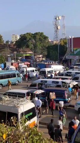 The video shows a busy street scene in an urban area with a view of a mountain in the background. There are numerous vans and minibuses clustered together, some with luggage racks on top. People are walking around and between the vehicles, some interacting with each other. A tall communications tower is visible, along with various buildings and storefronts. The sky is clear and the lighting suggests it's daytime. Overall, the scene depicts a bustling transportation hub with a scenic mountain backdrop.