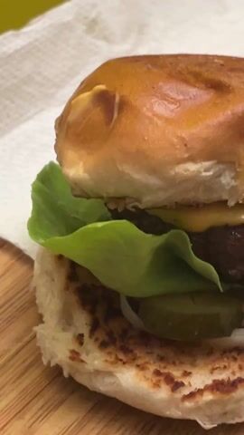 A close-up shot shows a hamburger on a wooden board. A paper napkin is positioned beneath the burger. The burger contains a browned bun, a beef patty, a slice of cheese that is melting, lettuce, sliced pickles, and sliced onions. Another burger can be seen slightly out of focus in the background.