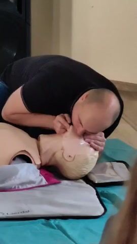 A man in a black polo shirt and blue jeans kneels on the floor next to a CPR training mannequin. He is demonstrating how to perform CPR. The mannequin is lying on its back on a blue cloth, with a gray and pink mat underneath its head and shoulders. The man first tilts the mannequin's head back and opens its airway. Then, he places his hands on the mannequin's chest and begins chest compressions. The scene is indoors, with a light-colored wall and a wooden floor visible in the background.