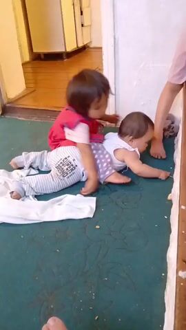 The video shows two young children playing on a dark green carpet in a room. The older child, wearing a red top and purple polka dot pants, is sitting on the back of the younger child, who is lying on their stomach. The younger child is wearing a white onesie with stripes and polka dots. Both children appear to be babies or toddlers. 

In the background, a wooden floor and a white cabinet are visible, suggesting a domestic setting, possibly a kitchen or hallway. A person's legs, wearing pink shorts, are visible on the right side of the frame, indicating an adult is present. The lighting suggests it is daytime. The children appear engaged in play and interaction. The scene is likely taking place indoors in Kotormo, Kyrgyzstan.

On the younger child's onesie, the words "SUPER BRAND" are visible in text.