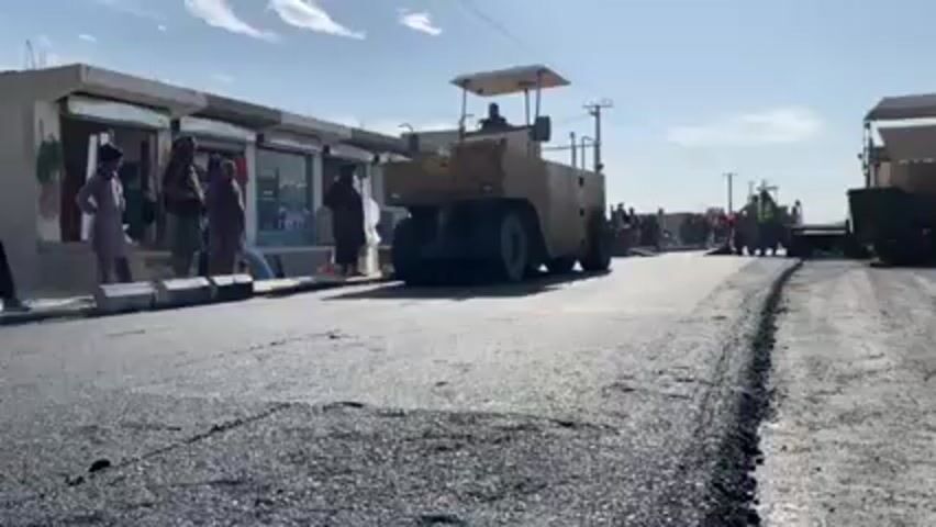The scene depicts road paving in what appears to be a small town or village setting. On the left side of the frame, a row of small shops or businesses can be seen, with several men standing in front of them, observing the work. In the middle of the street, a large road roller is slowly moving away from the camera, compacting freshly laid asphalt. There is another piece of road construction equipment further down the road, with several construction workers in the vicinity. The day is sunny, and the sky is mostly clear with some scattered clouds. The overall scene suggests a road construction project in progress in a populated area.