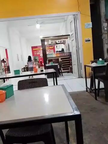 The video shows the interior of a casual eatery, possibly a restaurant or food stall, located in Meunasah Pupu, Indonesia. The setting appears to be indoors, with open-air elements suggesting a warm climate. The time of day is unclear, but the lighting suggests it could be evening or a dimly lit interior.

In the foreground, there are two tables set with chairs. On one table, there is a bottle of red sauce, a plastic container with an orange lid, and a green dispenser. The tables have a white surface with black metal frames. The floor is a gray, concrete-like material.

Further into the eatery, there are more tables and chairs. The walls are painted white, and the ceiling has exposed beams and lighting fixtures. A sign is visible on the back wall, with Indonesian text that reads "Bakso Kadir Mad". This likely indicates the name of the establishment or a type of food served.

Two individuals are the main subjects. In the background, a woman and a child are visible, seemingly engaged in activity within the eatery. In the mid-ground, a man wearing glasses and a blue polo shirt is seated, looking down at and interacting with a smartphone. His posture suggests he is relaxed.

The overall atmosphere seems to be relaxed and casual, typical of a local eatery. There are no clear indications of weather conditions. The presence of the smartphone suggests modern life integrated into the everyday setting.