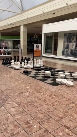 A wide shot shows the interior of a brightly lit shopping mall with a high, arched ceiling and a tiled floor. In the foreground, a large outdoor chessboard is set up, complete with oversized chess pieces and checkers. The board is situated on a black and white checkered mat. To the right of the chess set, a display case with various candies is visible, and further down the hall, a JCPenney store entrance is apparent. A digital display stands near the candy display. The mall appears mostly empty, with a few people visible in the distance near the JCPenney entrance. The lighting is consistent throughout, indicating daytime.