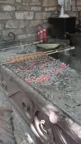 The video shows two skewers of raw ground meat, likely kebabs, being grilled over hot coals. The coals are glowing red and covered in ash. The grill appears to be made of metal and is situated in front of a stone wall. In the background, there is a fire extinguisher and a fan. The meat is positioned horizontally on the skewers, resting just above the coals. The camera angle is from a slightly elevated position looking down at the grill.