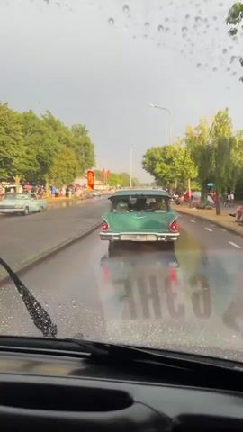 The video shows a view from inside a vehicle, looking out through the windshield which is wet with rain and has windshield wipers. The vehicle is on a wet road, suggesting it is raining or has recently rained. In front of the vehicle, there are several classic cars driving on the road. On the sides of the road, there are trees and sidewalks, and many people are gathered, some standing and some sitting. There appear to be tents or stalls set up along the side of the road, possibly for a street fair or car show. The overall atmosphere seems to be of an outdoor event taking place despite the weather.