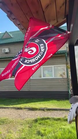 The video shows a red flag hanging from a wooden structure. The flag has a circular logo in the center with text in a Cyrillic script. The background includes a house with green roofing, a green lawn, and a clear blue sky. The flag is swaying in the wind.