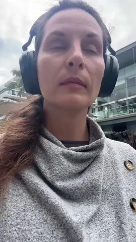 The video shows a close-up, medium shot of a woman with brown hair wearing large black headphones and a grey sweater with buttons. She is outdoors, and the background appears to be a street with buildings and some greenery. The woman's expression is neutral, and she seems to be looking forward. The video is shaky, suggesting it may have been filmed by the woman herself.