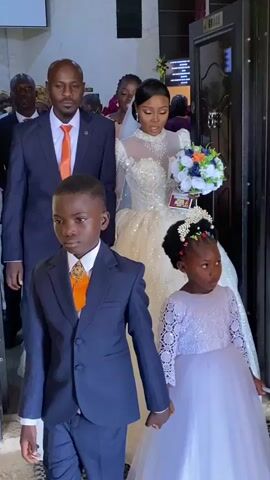 This video shows a wedding procession. In the foreground, two children, a boy and a girl, are walking hand-in-hand. The boy is wearing a navy blue suit with an orange tie and appears to be a ring bearer. The girl, likely a flower girl, is in a white lace dress with a flower crown. Behind them, a man and woman, presumably the groom and bride, are walking. The man is in a navy blue suit with an orange tie, and the bride is in a white wedding gown with a long train and is holding a bouquet. They are followed by other wedding guests. The scene appears to be indoors, possibly at a church or reception venue, with a doorway visible in the background. The overall atmosphere seems formal and celebratory.