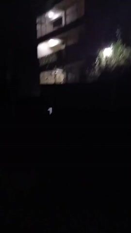 The video begins focusing on a three-story building at night. Lights are on in the units. The camera then pans to the right, away from the building, and the scene becomes very dark with only a single white streak of light visible.