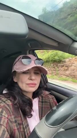 The video shows a woman driving a car down a road with stone walls and buildings visible through the window. She is wearing a pink cap with sunglasses on the brim, a pink hoodie, and a brown and red plaid jacket. The music playing in the car is upbeat and dance-oriented. She looks at the camera and smiles, waving her hand.