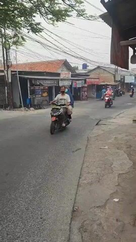 The video shows a street scene with several motorcycles. In the foreground, two people ride a red and black motorcycle. In the background, other people on motorcycles are seen. The street is lined with shops and buildings. Overhead, numerous electrical wires are visible. The sky is overcast.