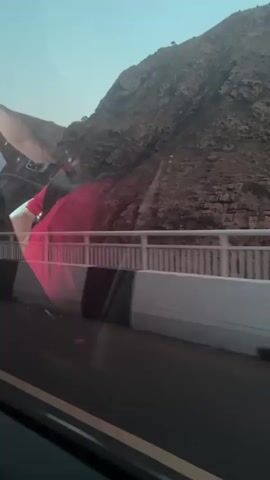 The video shows a view from inside a vehicle. A person wearing a red garment is visible, partially obscuring the view of a mountainous landscape and a bridge. The bridge has white railings and black and white concrete barriers. The road surface is visible in the foreground. The sky is clear and the lighting suggests daylight. The vehicle appears to be moving, as indicated by the motion blur in the footage.