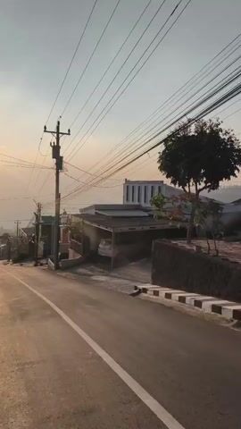 The video shows a residential street in an urban or semi-urban area during what appears to be either sunrise or sunset, given the warm, golden light. The street is paved and has a white line marking the center. On the right side of the street, there are houses, some with carports. A white car is visible under a carport. There are numerous utility poles with a dense network of electrical wires stretching across the scene. The buildings are of varying designs and colors, with some featuring modern architectural elements. On the left side, more houses and some trees are visible, and the street slopes downwards. The sky is clear with a hazy glow, and the sun is partially visible, casting long shadows. There are no people visible in the video.