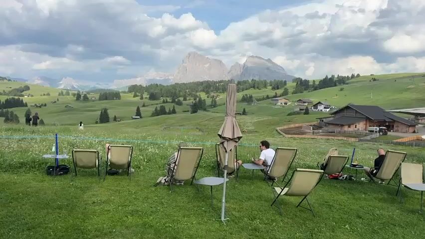 This media file captures a serene outdoor scene with several people relaxing on chairs in a lush green meadow. The setting is a picturesque mountain landscape, with rolling hills dotted with trees and small buildings. In the background, imposing mountains rise majestically under a cloudy sky, suggesting a cool, possibly temperate climate. A large building, possibly a farm or restaurant, is visible on the right side of the frame.

The subjects are a group of individuals seated in lounge chairs, some facing the mountains, others engaged in conversation or looking at their phones. An umbrella stands closed in the center foreground, a silent sentinel. The overall atmosphere is one of tranquility and leisure, with people enjoying the natural beauty of the location.

The time of day appears to be afternoon, given the lighting conditions. The weather is cloudy but does not appear to be raining. The location cues, such as the alpine scenery and architecture, strongly suggest the Dolomites region of Italy, specifically Kastelruth - Castelrotto, as indicated. There is no visible text within the frame. The emotions conveyed are peacefulness and relaxation, as people are comfortably seated and seemingly enjoying their surroundings.