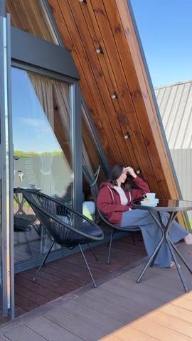 A young woman with dark hair sits on a chair on a wooden deck. She is wearing a maroon hooded sweatshirt and blue pants. She has her hand on her head and is looking away from the camera. On a small round table in front of her is a mug and a plate with a pastry. Next to her is another chair. Behind her is a modern A-frame building with large windows and wooden siding. The sky is blue and clear.