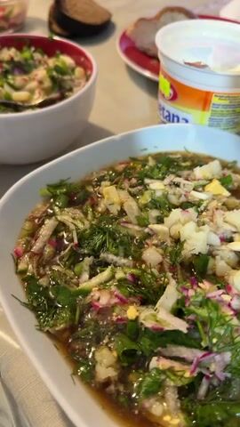 A close-up, vertical shot focuses on two bowls of soup and a bottle of beer. The bowl in the foreground is white and oval-shaped, filled with a dark broth containing chopped vegetables, herbs, and pieces of egg and potato. A dark liquid, presumably beer, is being poured from a brown glass bottle into the soup. The bottle's neck and cap are visible at the right edge of the frame. In the background, another white bowl with a red rim contains a similar soup, with a spoon resting in it. To the right of the bowls, a white plastic container with a colorful label is visible, likely containing sour cream or yogurt, and a slice of dark bread. The setting appears to be a table, with a light-colored tablecloth. The lighting is natural, suggesting an indoor environment. There is no visible interaction between people, only the action of preparing the soup.