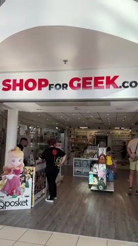 The video shows the entrance to a store called "SHOPforGEEK.COM". Inside, there are shelves filled with merchandise, likely collectibles and merchandise for geeks. A man wearing a beige t-shirt and yellow shorts is standing in the middle of the store, looking at the shelves. He is holding a beige baseball cap and has a small bag slung over his shoulder. Another person, wearing a black t-shirt and black pants, is standing behind a counter on the left side of the store, interacting with a customer who is partially visible. There are displays of merchandise at the entrance, including what appears to be a character figurine and posters. The store has a tiled floor and is brightly lit with overhead lights.