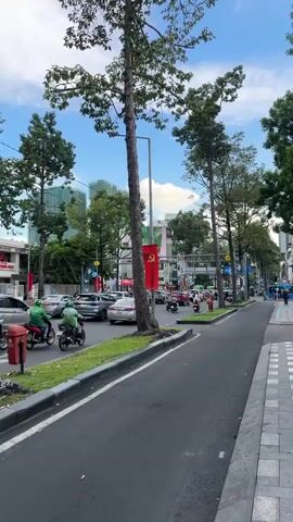 The video shows a street scene in a city, likely in Southeast Asia, with heavy traffic, mostly motorcycles. Tall trees line the street, casting shadows on the road. A red flag with a yellow star, characteristic of communist flags, is prominently displayed on a lamppost. Cars and motorcycles navigate the busy intersection, with some motorcycles appearing to be delivery vehicles. Pedestrians can be seen on the sidewalk in the distance. The sky is blue with some clouds, suggesting it is daytime. The overall atmosphere appears to be one of bustling urban activity.