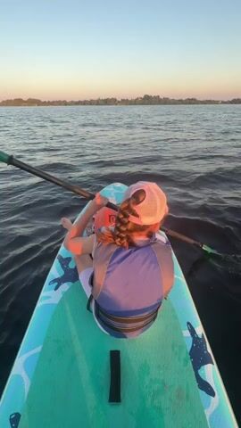 A person is sitting on a paddleboard in the water. They are wearing a life jacket and a hat, and their hair is in a braid. They are holding a paddle. The sun is setting in the background, casting a warm glow over the scene. The water is calm, with gentle ripples. The paddleboard is decorated with a blue and white pattern.