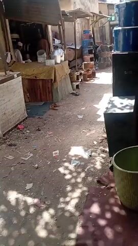 The video shows a bustling alleyway, with a man walking while carrying a metal bucket. The alley is narrow and cluttered with various items, including metal drums, buckets, and scraps of paper and trash scattered on the ground. Some people are seen sitting and working. The alley is shaded in some areas, with bright sunlight in others. The camera follows the man with the bucket as he walks, providing a view of the surroundings. The sounds of work and activity can be heard in the background.