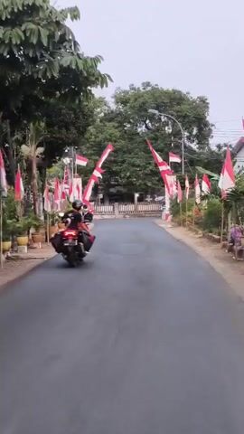 A person rides a motorcycle down a paved road lined with trees and houses decorated with Indonesian flags. The flags are red and white and are mounted on poles and form arches over the road. The sky is overcast.