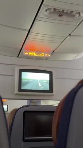From an airplane, a video screen shows the runway ahead. The runway stretches into the distance, marked with white lines and runway lights. Above the screen are overhead panels with safety indicators lit in orange, including a seatbelt reminder. A person is partially visible in the lower right, suggesting the video is taken from a passenger's perspective.