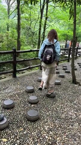Several people are walking on a path consisting of rounded, gray stones, with similarly colored stepping stones embedded among them. The area is wooded, with trees visible in the background.  The ground around the stones is covered in tiny, loose pebbles. The people are dressed casually in shorts, skirts, long pants and shirts. All of the people are wearing backpacks.